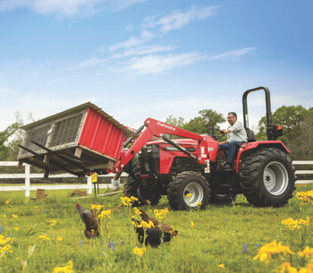 Red Mahindra tractor with loader lifting a barn structure in a field with chickens and flowers.
